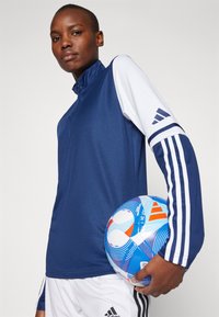 Marineblå atletisk lynlås bluse med hvide ærmer og tre hvide striber. Modellen holder en multifarvet fodbold med bølgende mønstre.