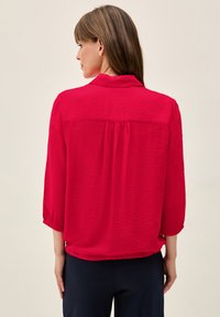 Blouse rouge texturée avec un col et des manches trois-quarts. Présente un dos avec empiècement et un détail froncé pour une coupe décontractée.