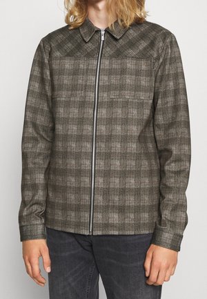 Personne portant une veste zippée à carreaux vert brun avec col et manches longues, associée à un pantalon gris foncé.