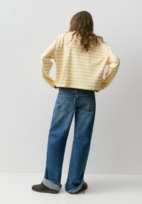 Personne aux cheveux longs, portant une chemise à manches longues rayée jaune et blanche et un jean bleu avec des clous en forme d'étoile sur les poches arrière, debout les mains sur les hanches.