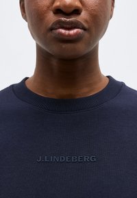 Κοντινό πλάνο ενός ατόμου που φοράει μπλε σκούρο φούτερ με το λογότυπο "J.LINDEBERG" ανάγλυφο στο στήθος, με απλό φόντο.