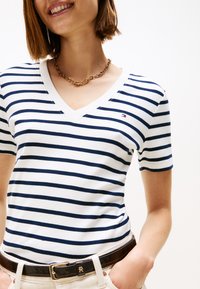 Smaidīga persona, kas valkā baltu un tumši zilu svītrainu V veida kakla T-kreklu, zelta ķēdes kaklarotu un tumši brūnu jostu ar zelta sprādzi.