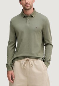 Polo a maniche lunghe in cotone verde oliva. Presenta un colletto piatto, una patta con bottoni e un piccolo logo sul petto. Abbinata a pantaloncini beige con coulisse.