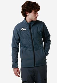 Giacca grigia scura con colletto alto, chiusura con zip e logo Kappa sulla spalla sinistra. Presenta pannelli testurizzati e tasche con zip.