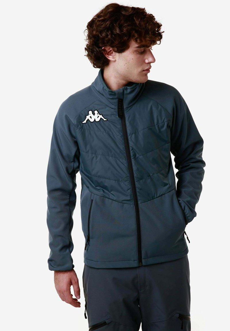 Giacca grigia scura con colletto alto, chiusura con zip e logo Kappa sulla spalla sinistra. Presenta pannelli testurizzati e tasche con zip.