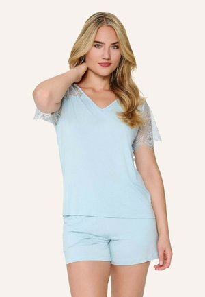 Lyshåret kvinde iført lyseblå kortærmet top med blonde detaljer og matchende shorts, med den ene hånd berørende sin nakke.
