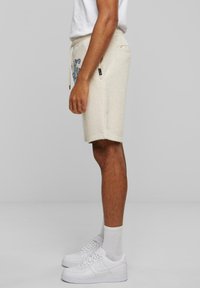 Shorts en coton beige avec taille à cordon et poches latérales, ornés d'une impression graphique bleue. Portés avec des baskets blanches et des chaussettes.