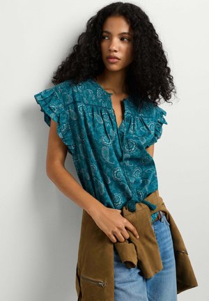 Frau mit lockigem Haar, die eine türkis gemusterte Bluse und blaue Jeans trägt und eine braune Jacke um die Taille gebunden hält.