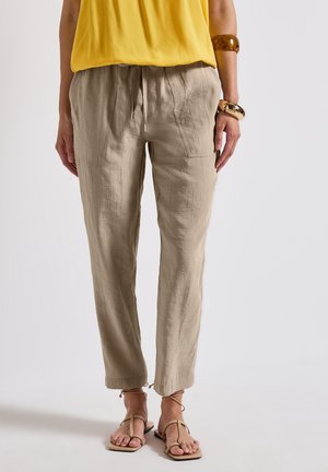 Kobieta ubrana w beżowe lniane spodnie ze sznurkiem, żółty top bez rękawów, jasnobrązowe sandały na paski i masywne bransoletki na jednym nadgarstku, stojąca na białym tle.