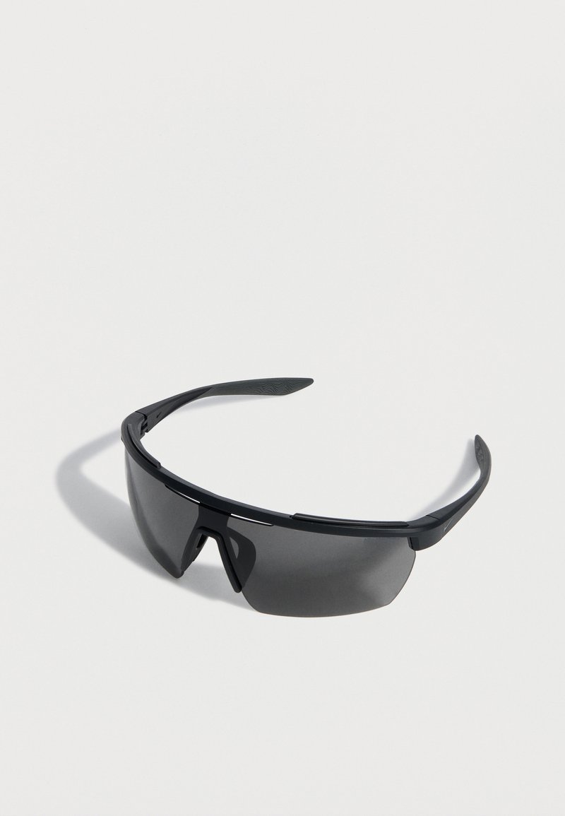 Μαύρα σπορ γυαλιά ηλίου με σκούρους, καμπυλωτούς φακούς και λεπτά πλαίσια, τοποθετημένα σε επίπεδη λευκή επιφάνεια.