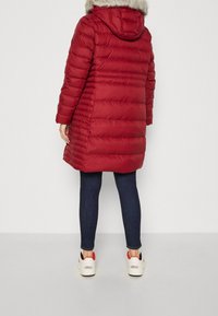 Persoon in een knielange rode pufferjack met een bontkap, donkere spijkerbroek en witte Adidas sneakers, staande op een effen achtergrond.