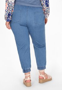 Vaaleansiniset puuvillaiset jogger-housut, joissa on elastiset hihnat ja vyötärö. Kaksi takataskua näkyvissä. Yhdistetty beigeihin espadrille-sandaaleihin.