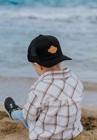 Mustaline trucker-kate, millel on pruun nahkplekk, valge ruuduline flanellist särk, heledad sinised teksad ja mustad tossud, istub liivases rannas.