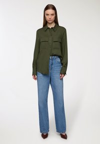 Camicia a maniche lunghe verde oliva con bottoni e due taschini sul petto, abbinata a jeans blu a vita alta e dalla vestibilità larga, e scarpe marroni a punta.