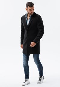  Schwarzer Wollmantel mit tailliertem Design, einreihigem Knopfverschluss und Seitentaschen, kombiniert mit blauen Jeans und weißen Sneakers.