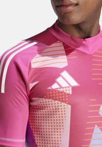 T-shirt de sport rose en tissu texturé, avec des motifs géométriques dans des tons d'orange et de blanc, et trois rayures blanches sur l'épaule.