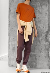 Roest oranje T-shirt met een logo, crèmekleurige trui gestrikt in de taille, paarse joggingbroek en witte sneakers met oranje en zwarte accenten.