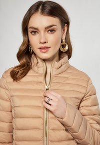 Jachetă puffer bej cu cusături orizontale, guler înalt și fermoar aurit. Modelul poartă cercei mari și o verigă din argint. Textură netedă.