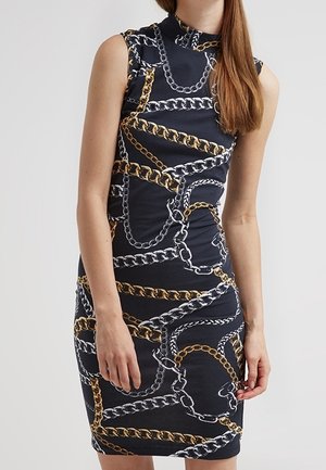Femme portant une robe noire sans manches avec un imprimé de chaînes dorées et argentées, debout devant un fond clair uni.