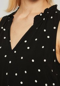 Blouse noire à pois blancs, encolure en V et accents de volants aux épaules. Tissu doux à la texture lisse, design sans manches.