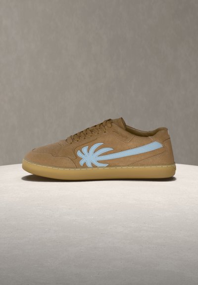 Καφέ σουέντ αθλητικό παπούτσι με φωτεινό γαλάζιο λογότυπο φοίνικα στο πλάι και σοκολατί σόλα, εκτεθειμένο σε μια λεία γκρίζα επιφάνεια με γκρίζο φόντο.