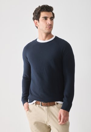 Suéter de punto en color azul marino oscuro con puños acanalados y un escote redondeado, superpuesto a una camiseta blanca de cuello redondo. Llevado con pantalones beige y un cinturón marrón.