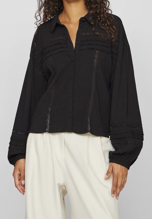 Kobieta w luźnej czarnej bluzce zapinanej na guziki z koronkowymi detalami i bufiastymi rękawami, w połączeniu z jasnobeżowymi spodniami z wysokim stanem.