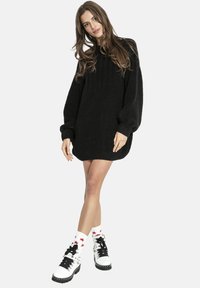 Czarna, oversize'owa sukienka swetrowa o prążkowanej fakturze, z długimi rękawami i zaokrąglonym dołem, zestawiona z białymi, sznurowanymi botkami oraz skarpetkami w gwiazdki.