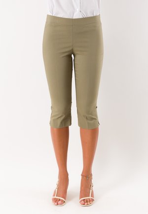 Donna che indossa pantaloni beige aderenti al ginocchio abbinati a sandali bianchi aperti con tacco alto e un braccialetto alla caviglia su uno sfondo semplice.