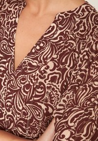 Burgundy mönstrad tyg med blommönster, som har en djup V-ringning och texturerade inslag i beige och metalltrådar.