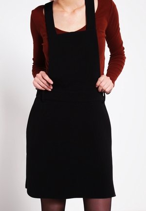 Person mit einem langärmligen braunen Oberteil unter einem schwarzen Trägerkleid, hält die Träger des Kleides, von den Schultern bis zu den Knien vor neutralem Hintergrund gezeigt.