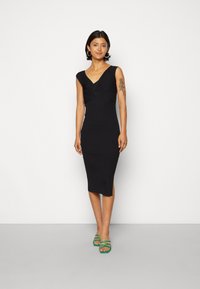 Vestido midi preto com textura canelada, decote em V e mangas curtas. Apresenta uma abertura lateral e uma silhueta justa. Combinado com sandálias verdes de tiras.