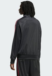 Černá sportovní bunda s vertikální žebrovanou texturou, vysokým límcem a červenými akcentovými pruhy podél ramen. Součástí jsou elastické manžety a lem.