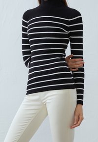 Suéter de cuello alto a rayas en blanco y negro con tejido acanalado, diseño ajustado, mangas largas, combinado con pantalones de color crema.