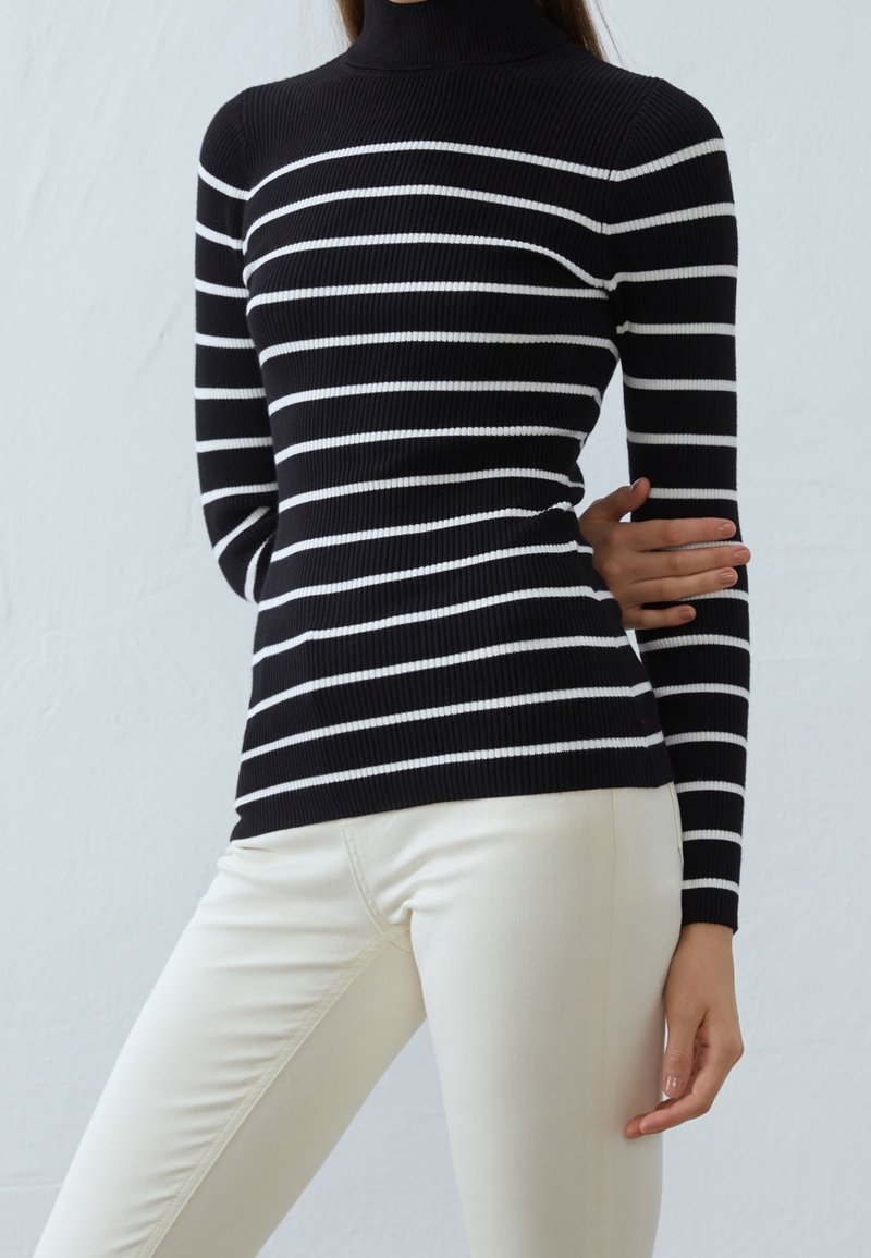 Suéter de cuello alto a rayas en blanco y negro con tejido acanalado, diseño ajustado, mangas largas, combinado con pantalones de color crema.