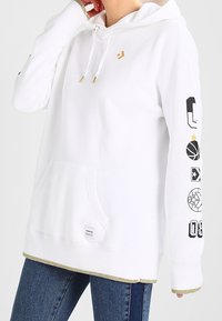 Haut à capuche blanc avec des embouts de cordon dorés, graphismes noirs et dorés sur le thème du sport sur la manche droite, poche avant, porté avec un jean bleu.