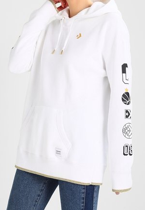 Felpa bianca con punte del cappuccio dorate, grafiche sportive nere e oro sulla manica destra, tasca frontale, indossata con jeans blu.