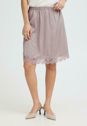 Donna con gonna mauve al ginocchio con orlo in pizzo, top bianco, tacchi slingback beige e anelli d'argento alle dita.