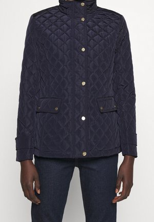 Gesteppte marineblaue Jacke mit Druckknöpfen, Klappentaschen und Stehkragen, getragen über dunklen Jeans, zeigt Torso und Arme des Modells.