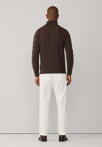 Suéter de punto trenzado marrón oscuro con cuello alto. Combinado con pantalones blancos y zapatos oscuros, mostrado por la parte de atrás. Textura suave, puños acanalados.