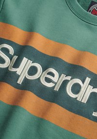 Zielona bluza z pomarańczowymi i ciemnozielonymi poziomymi paskami. Na piersi wyhaftowane w białym kolorze "Superdry". Gładka tekstura.