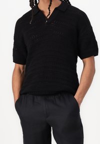 Czarny dzianinowy polo z fakturowanym wzorem, krótki rękaw i dekolt w serek, w zestawie z ciemnymi, luźnymi spodniami.