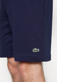 Short bleu marine porté sur une jambe, avec un petit logo crocodile vert près de l'ourlet sur le côté gauche.