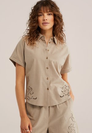 Femeie cu păr creț purtând o cămașă bej cu mânecă scurtă și nasturi, asortată cu pantaloni scurți, ambele având modele decorative decupate.