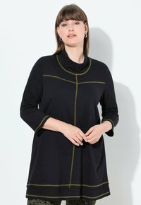 Vrouw met bruin haar, gekleed in een zwarte tuniek met lange mouwen en groene stikdetails, en een leggings met patroon, staand tegen een witte muur.