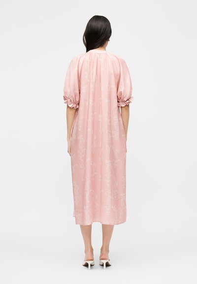 Γυναίκα με μακριά σκουρόχρωμα μαλλιά, που φοράει ένα άνετο, ροζ φόρεμα μέχρι το γόνατο με φουσκωτά μανίκια και άσπρα σανδάλια με τακούνι, να στέκεται μπροστά σε λευκό φόντο.