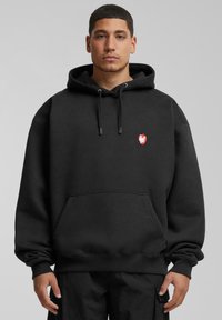 Schwarze Kapuzenjacke aus Fleece-Material, mit einer Fronttasche, Kordelzugkapuze und einem kleinen roten Logo auf der Brust. Gerippte Bündchen und Saum.