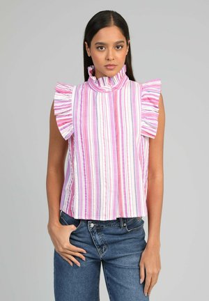 Femme portant un chemisier sans manches rayé rose, blanc et violet avec des épaules volantées et un col haut, associé à un jean bleu.