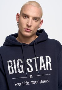 Tamsiai mėlynas džemperis su gobtuvu, pagamintas iš minkštos medžiagos, su dideliu baltu užrašu "BIG STAR" ir mažesniu šūkiu žemiau. Įtraukti reguliuojami raiščiai.