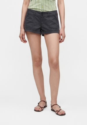 Person iført mørkegrå shorts, grøn ternet ærmeløs skjorte og brune flade sandaler med stropper, stående mod en ensfarvet baggrund.